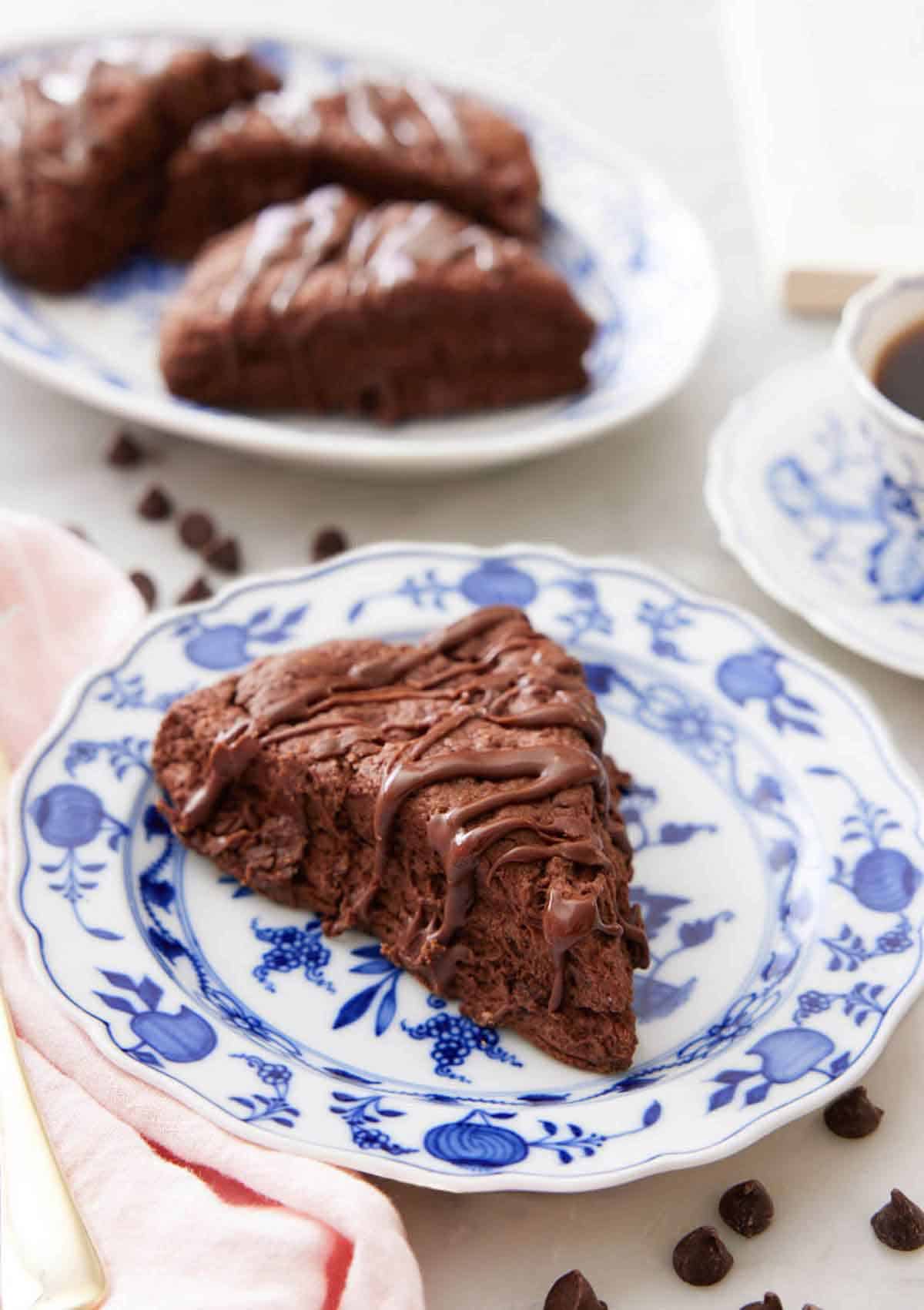 A plate with a chocolate scone with chocolate drizzled on top with a platter of scones in the background.