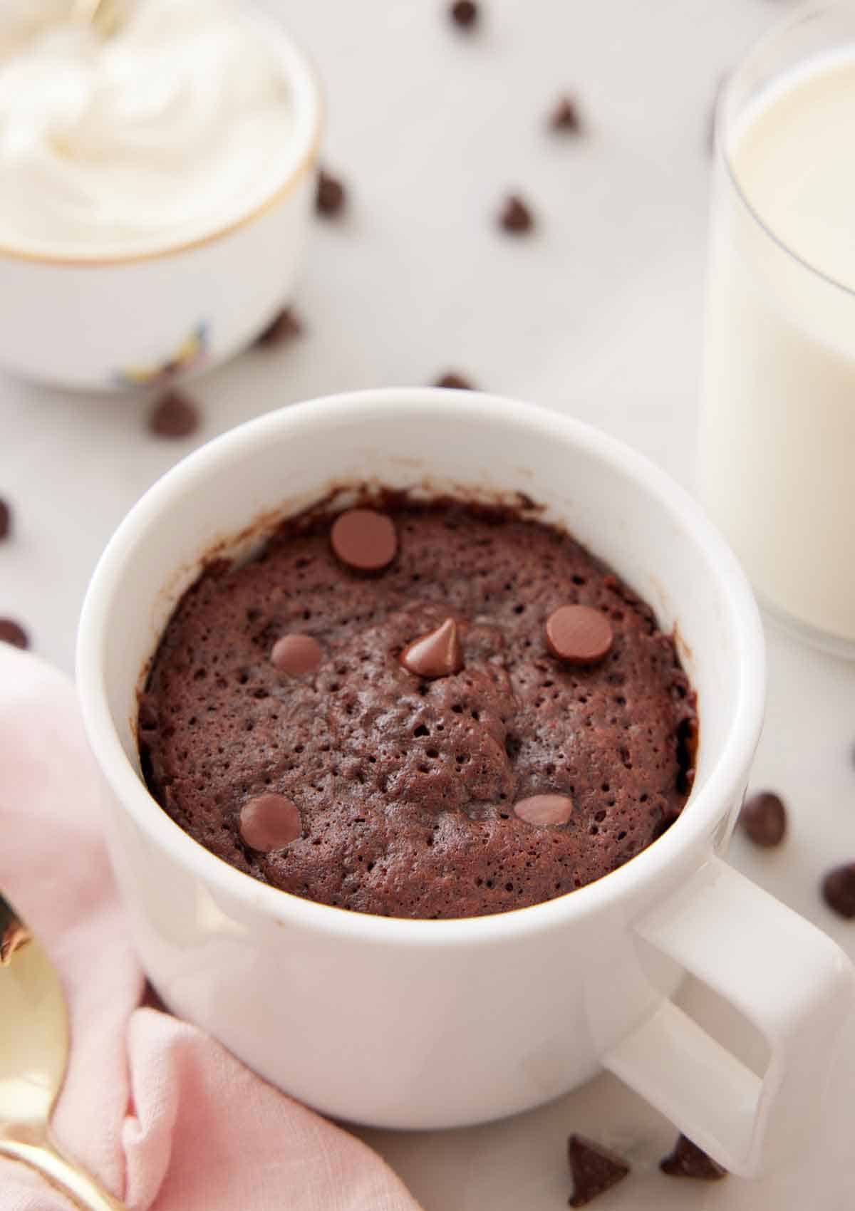 A mug cake with chocolate chips on top with more chocolate chips scattered in the back with a glass of milk.