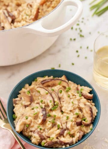 A bowl of mushroom risotto with chives garnished on top with a pot of more risotto and a glass of wine in the back.