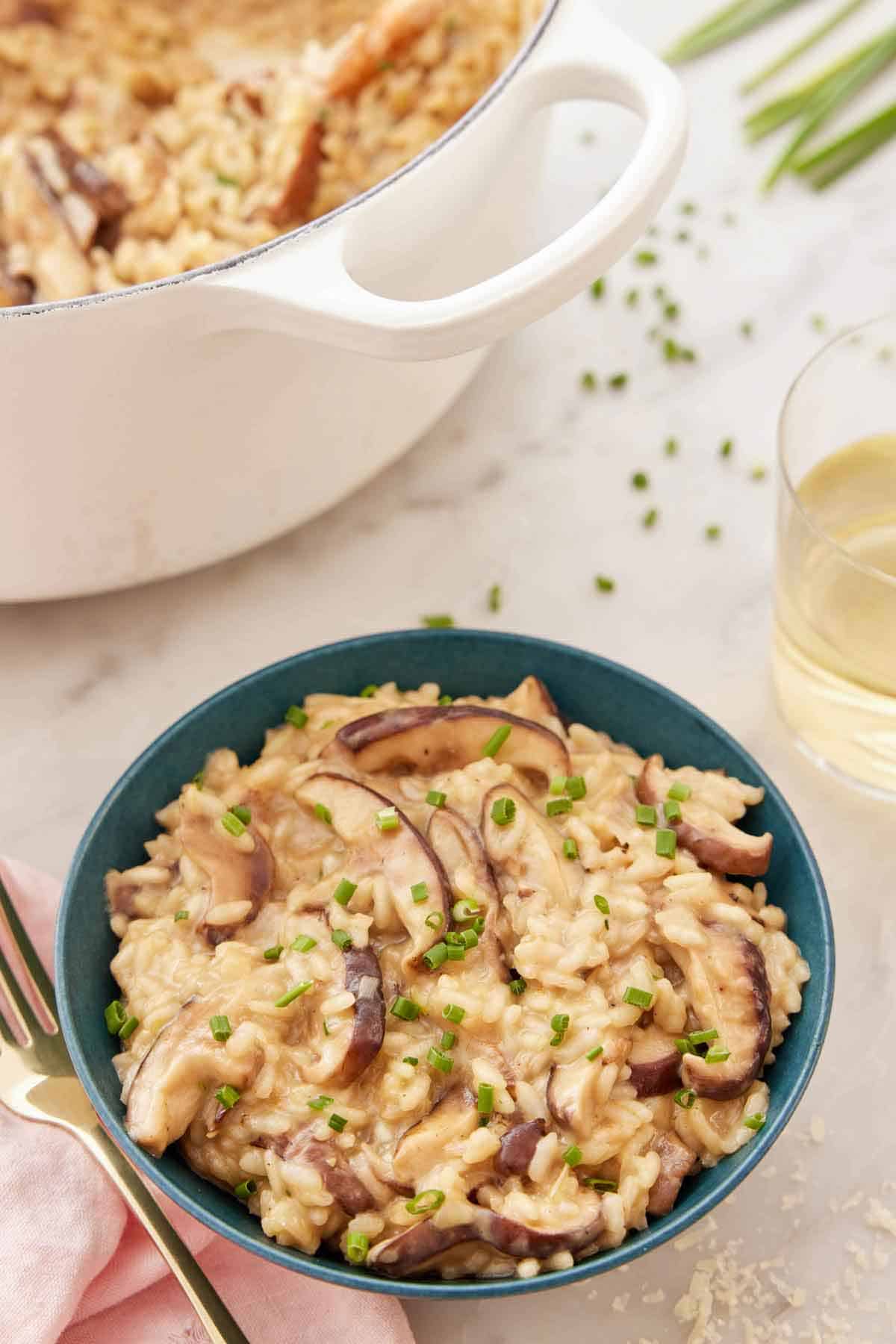 A bowl of mushroom risotto with chives garnished on top with a pot of more risotto and a glass of wine in the back.