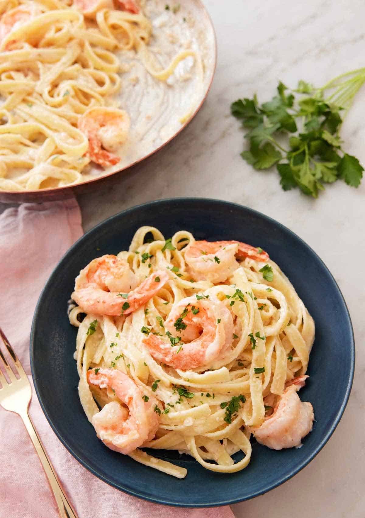 A plate of shrimp Alfredo with a bunch of parsley in the background along with a pan of the pasta.
