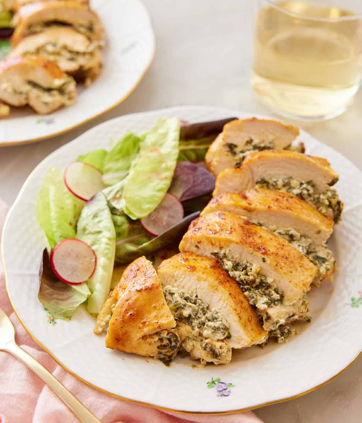 A plate with a spinach stuffed chicken breast, sliced, with salad on the side. A glass of wine and another plate of chicken in the background.