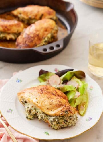 A plate with a spinach stuffed chicken breast with some salad with a cast iron with three more breasts in the background.