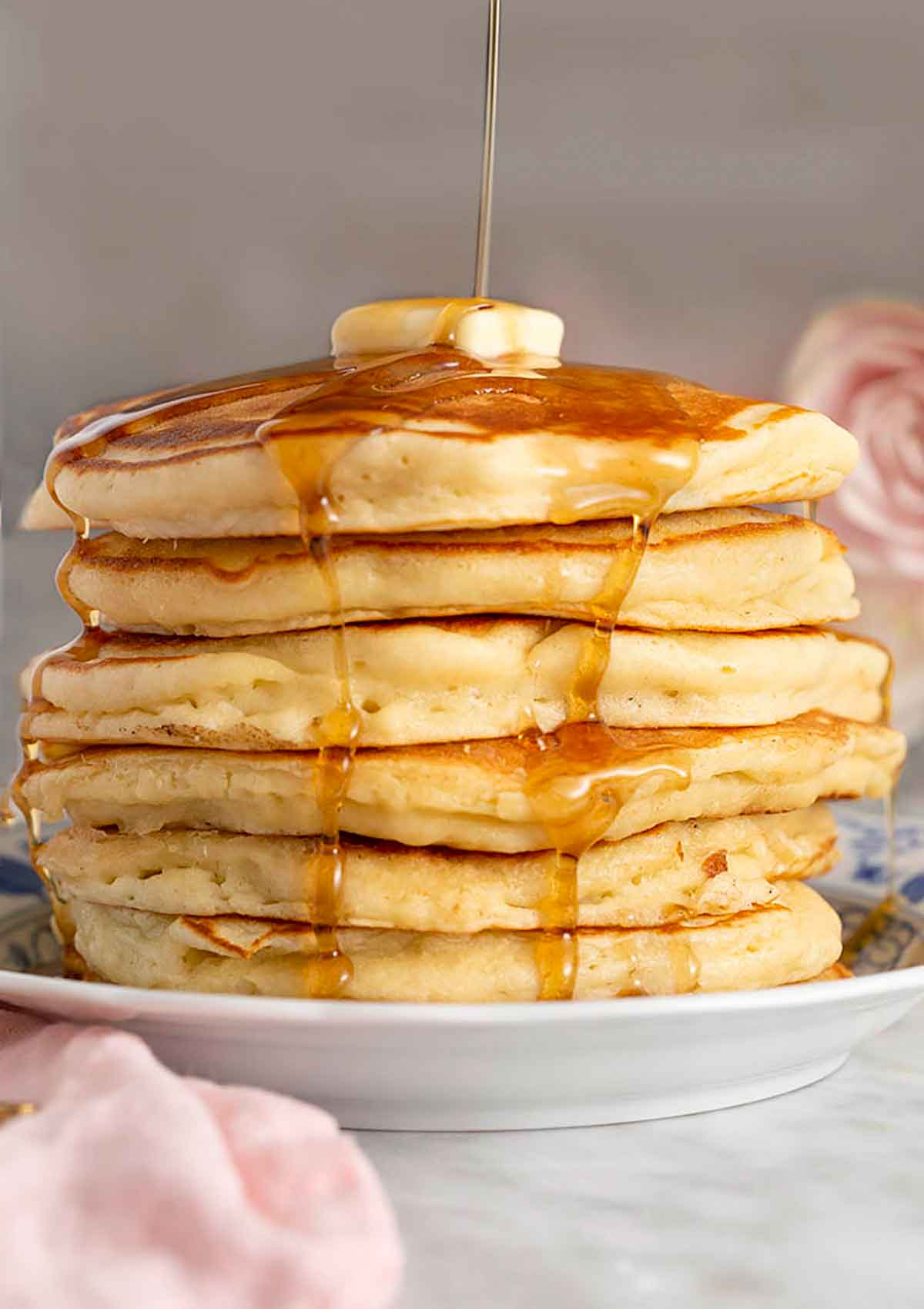 A tall stack of pancakes topped with a pat of butter with syrup being poured on top.