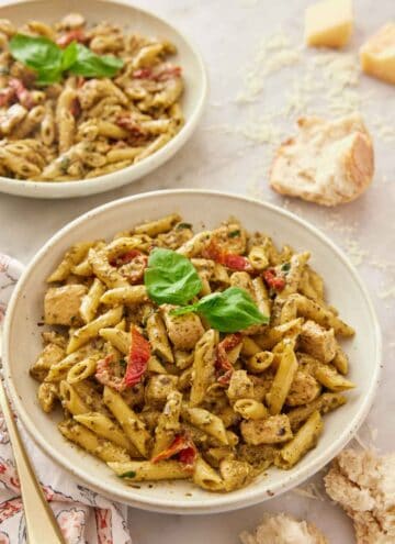 A plate of chicken pesto pasta with basil and sun-dried tomatoes with some bread, shredded parmesan and another plate of pasta in the background.