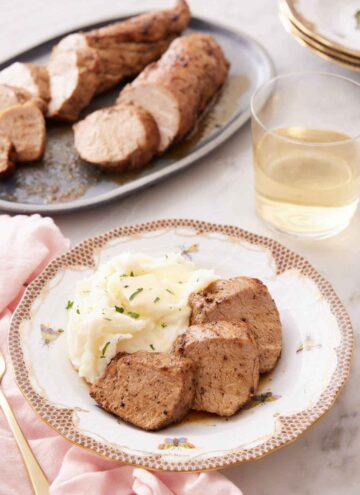 A plate with three pieces of pork tenderloin with mashed potatoes on a plate. A platter of more pork and a glass of wine in the background.