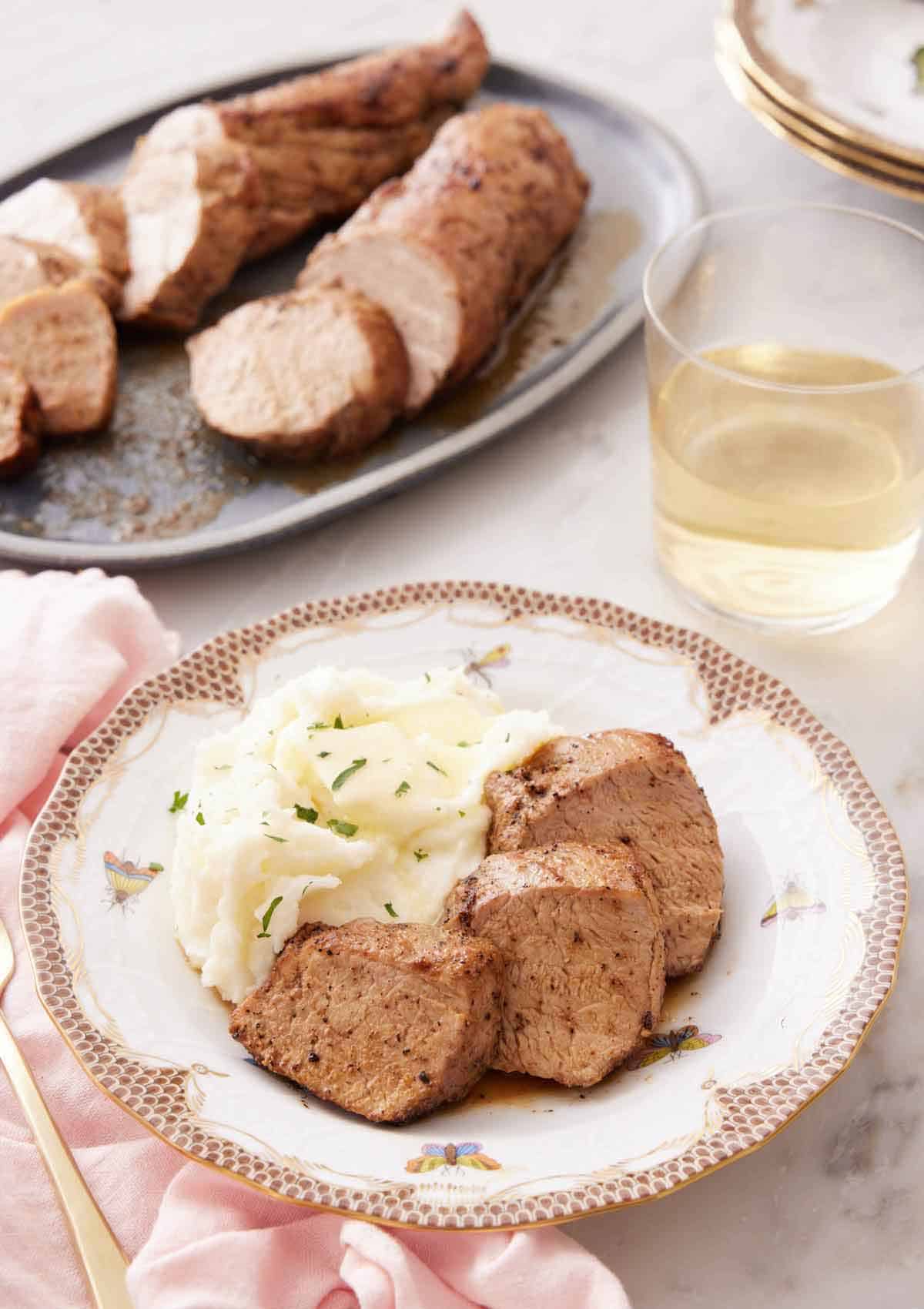 A plate with three pieces of pork tenderloin with mashed potatoes on a plate. A platter of more pork and a glass of wine in the background.