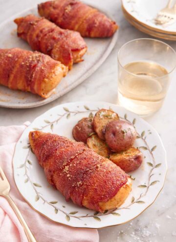 A plate with a bacon wrapped chicken breast with a side of baby potatoes. A glass of wine and platter of chicken in the back.