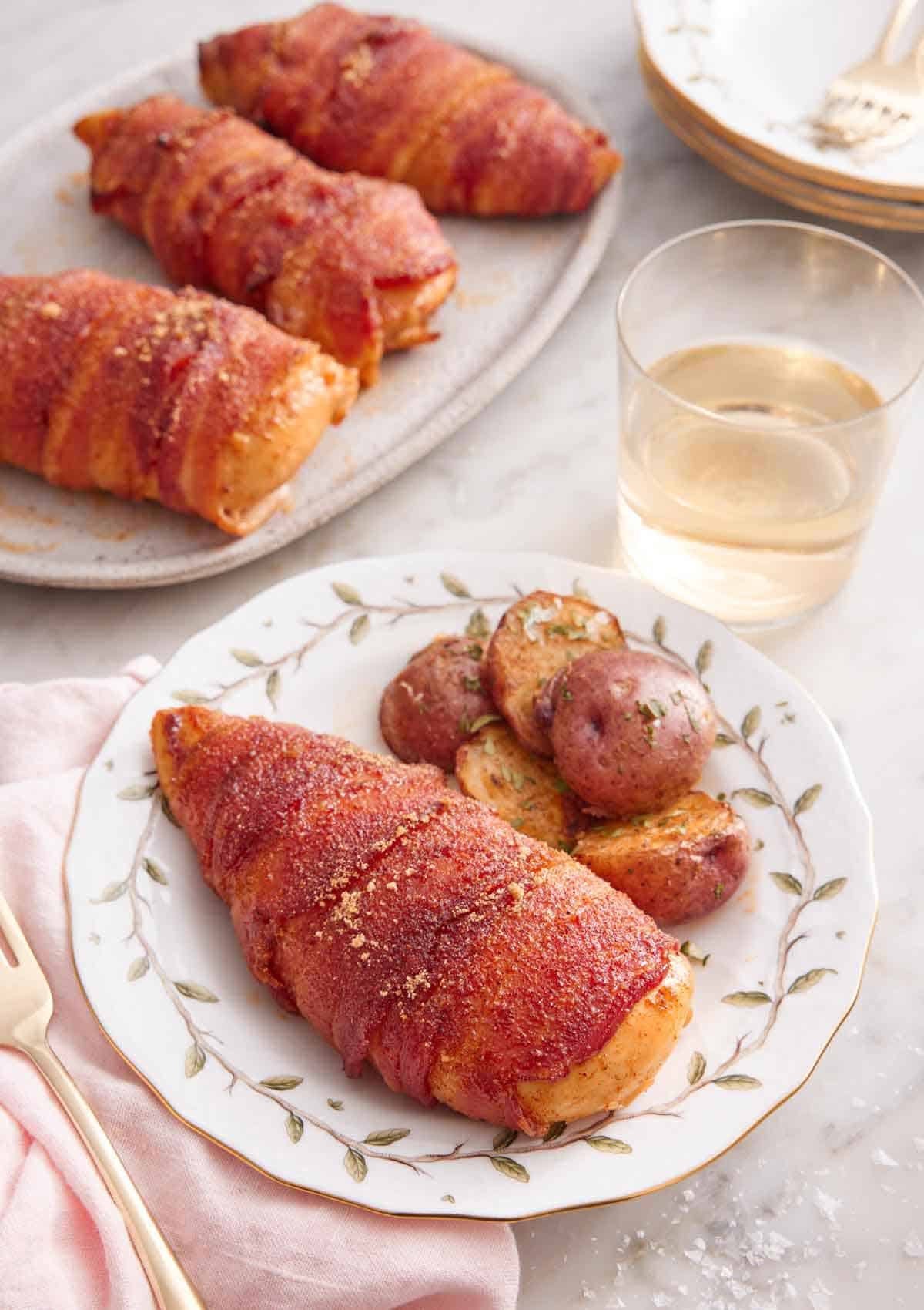 A plate with a bacon wrapped chicken breast with a side of baby potatoes. A glass of wine and platter of chicken in the back.