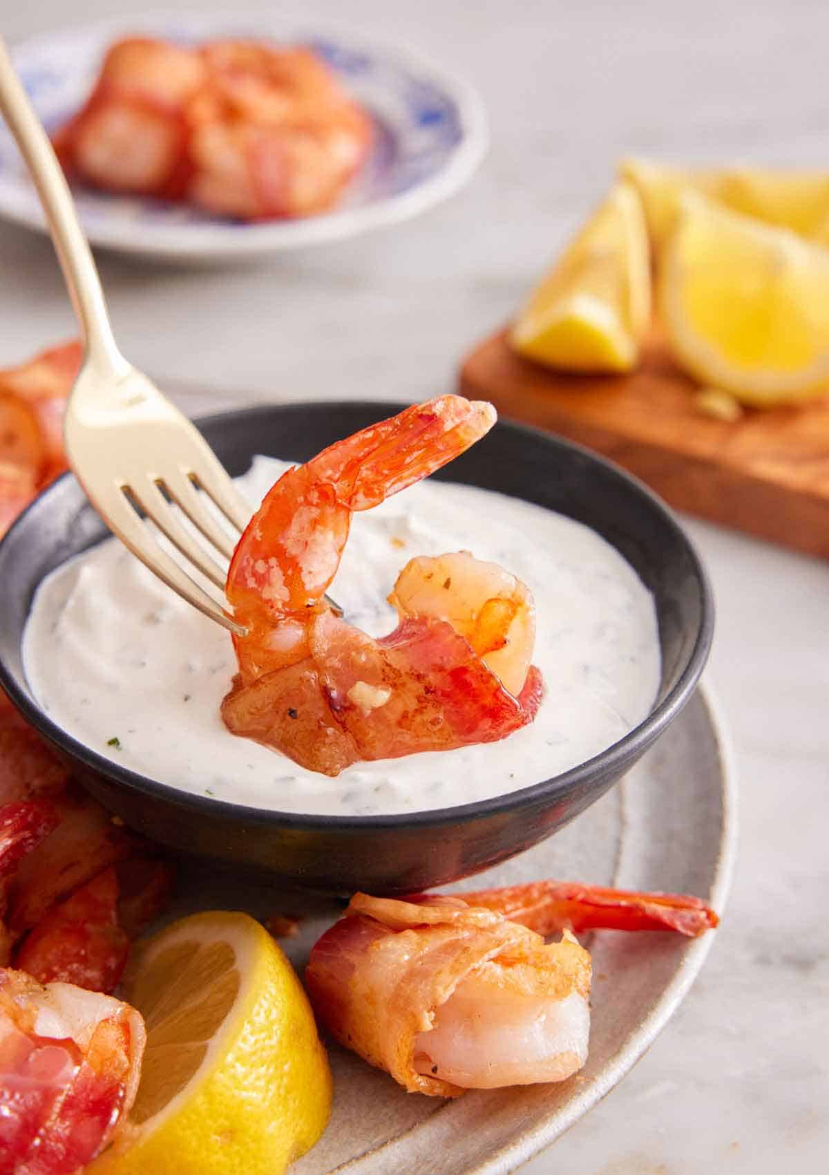 A bacon wrapped shrimp on a fork, dipped into white sauce.
