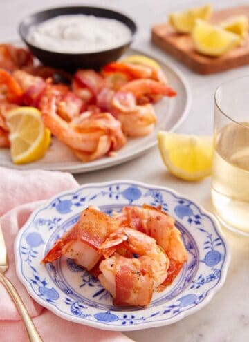 A plate with three pieces of bacon wrapped shrimp with more in the back with dip, lemons, and a glass of wine.