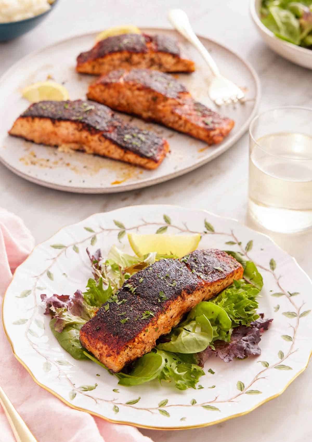 A plate with blackened salmon over salad with more salmon in the background.