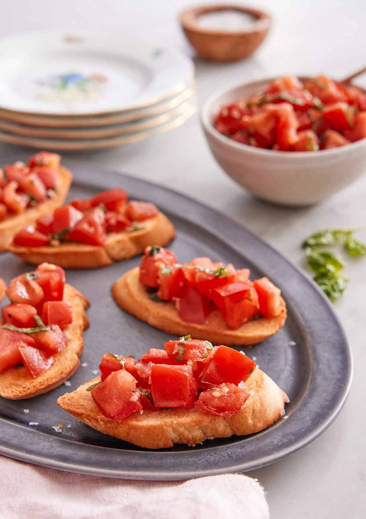 A platter with bruschetta on it with a bowl of tomato mixture in the back with a stack of plates.