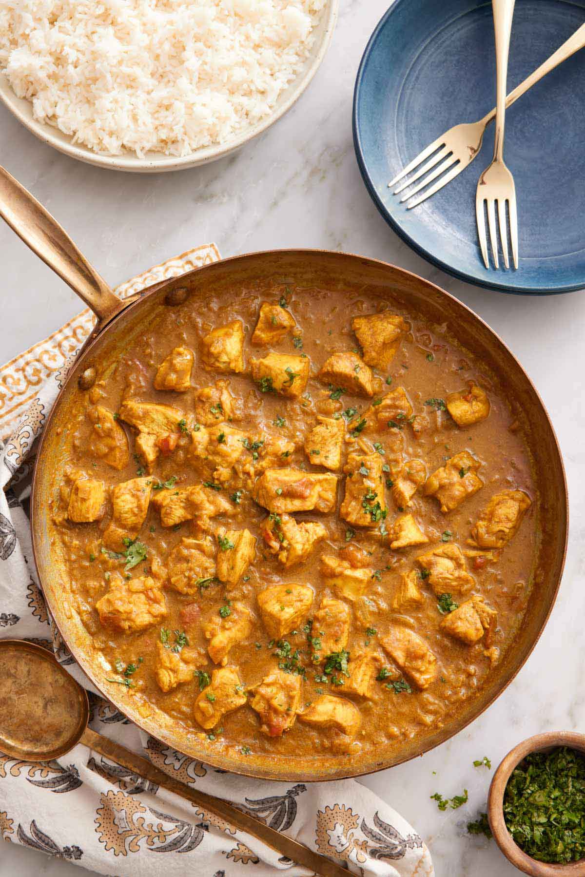 Overhead view of a skillet of chicken curry with a plate of rice off to the side with some forks and garnish.