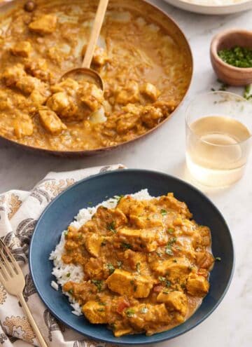 A bowl of chicken curry over rice with a skillet of more curry and glass of wine in the background.