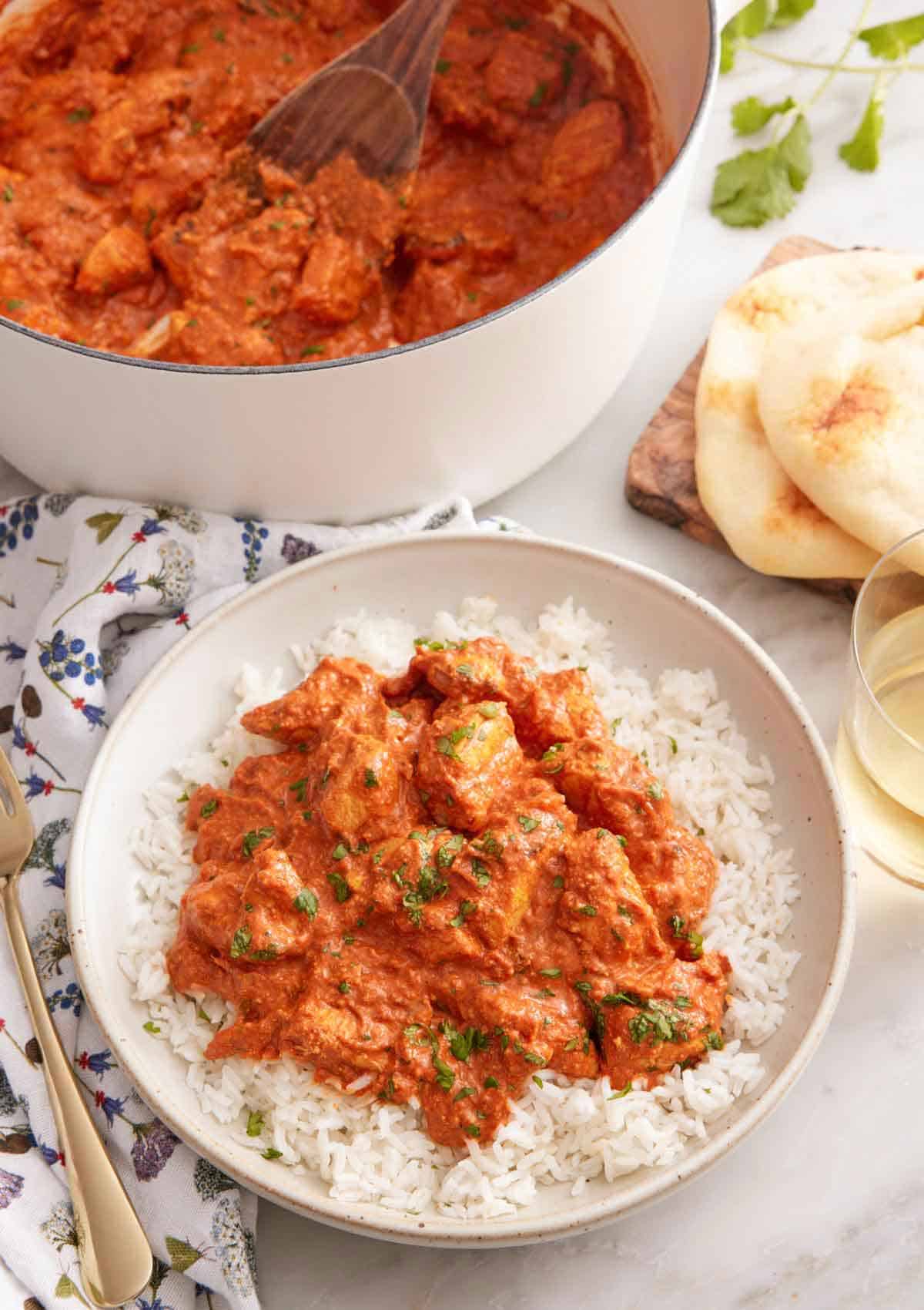 A bowl of chicken tikka masala over rice with a pot of more curry in the back along with naan.