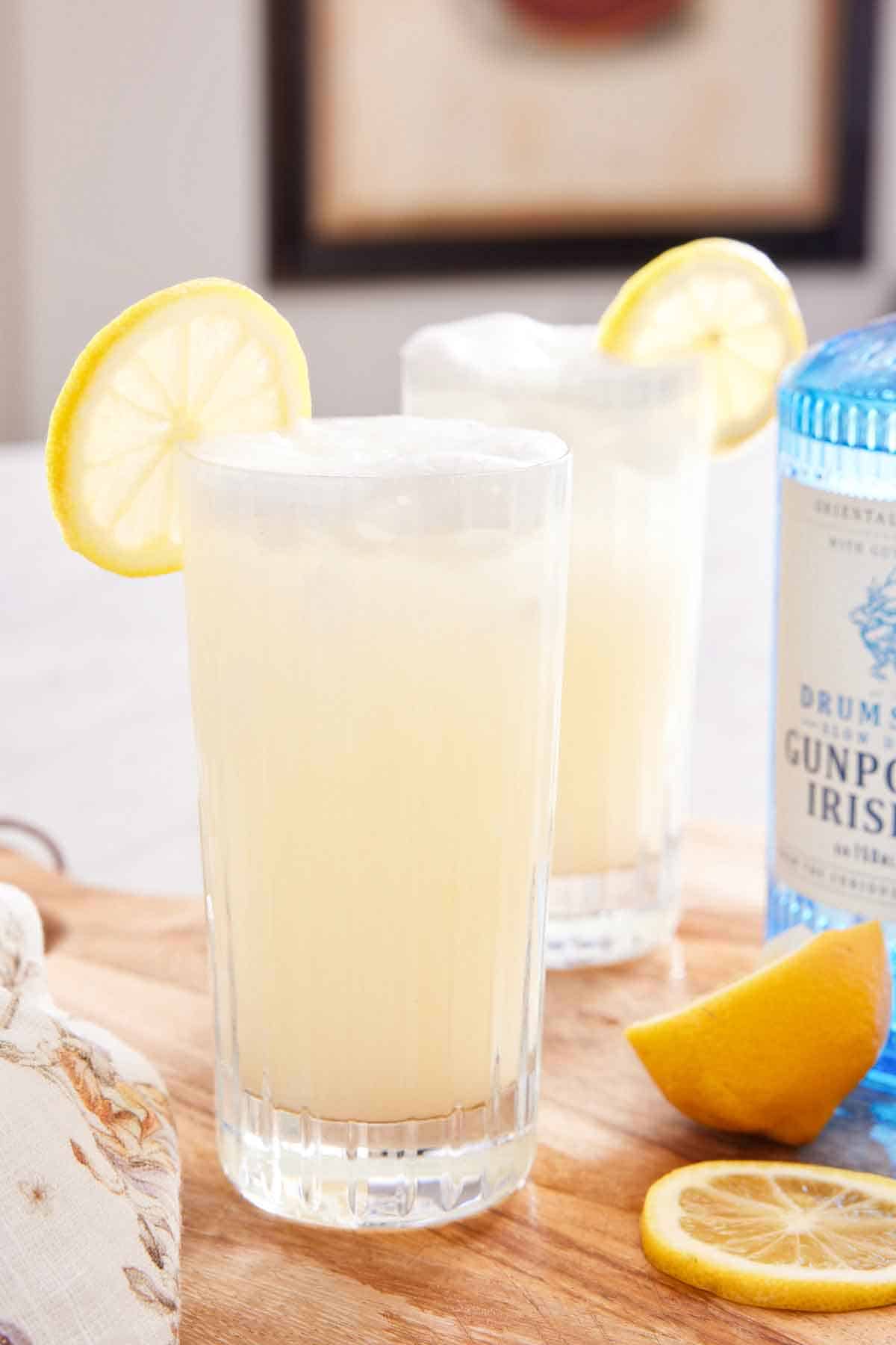A glass of Gin Fizz with a lemon slice garnish with an additional glass in the back with a bottle of gin.