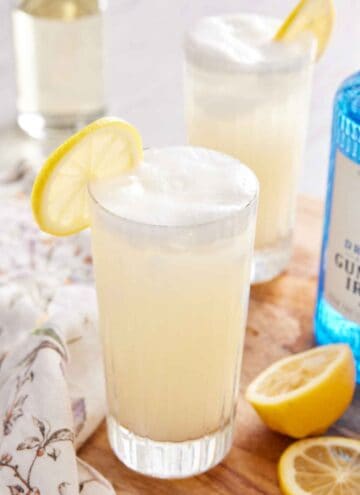 A tall glass of Gin Fizz with a lemon slice on the rim with another glass, a gin bottle, and cut lemons off to the side.
