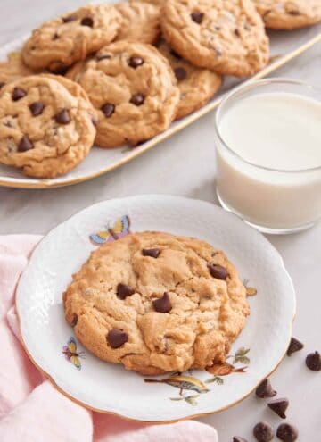 A plate with a peanut butter chocolate chip cookie with a platter of more cookies in the back along with a glass of milk.