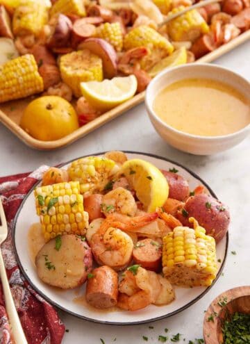 A plate of shrimp boil with a sheet pan and butter in a bowl in the back.