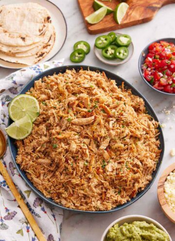 Overhead view of s platter of slow cooker carnitas with cut limes. Additional toppings such as guacamole, salsa, jalapenos on the side with some tortillas.