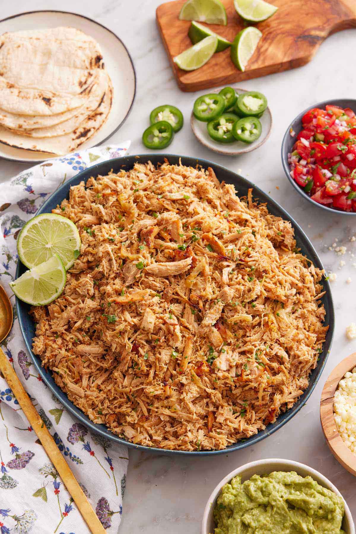 Overhead view of s platter of slow cooker carnitas with cut limes. Additional toppings such as guacamole, salsa, jalapenos on the side with some tortillas.