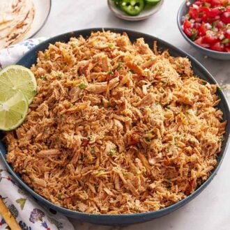 A large bowl of slow cooker carnitas with lime wedges. Some guacamole, salsa, and tortillas on the side.