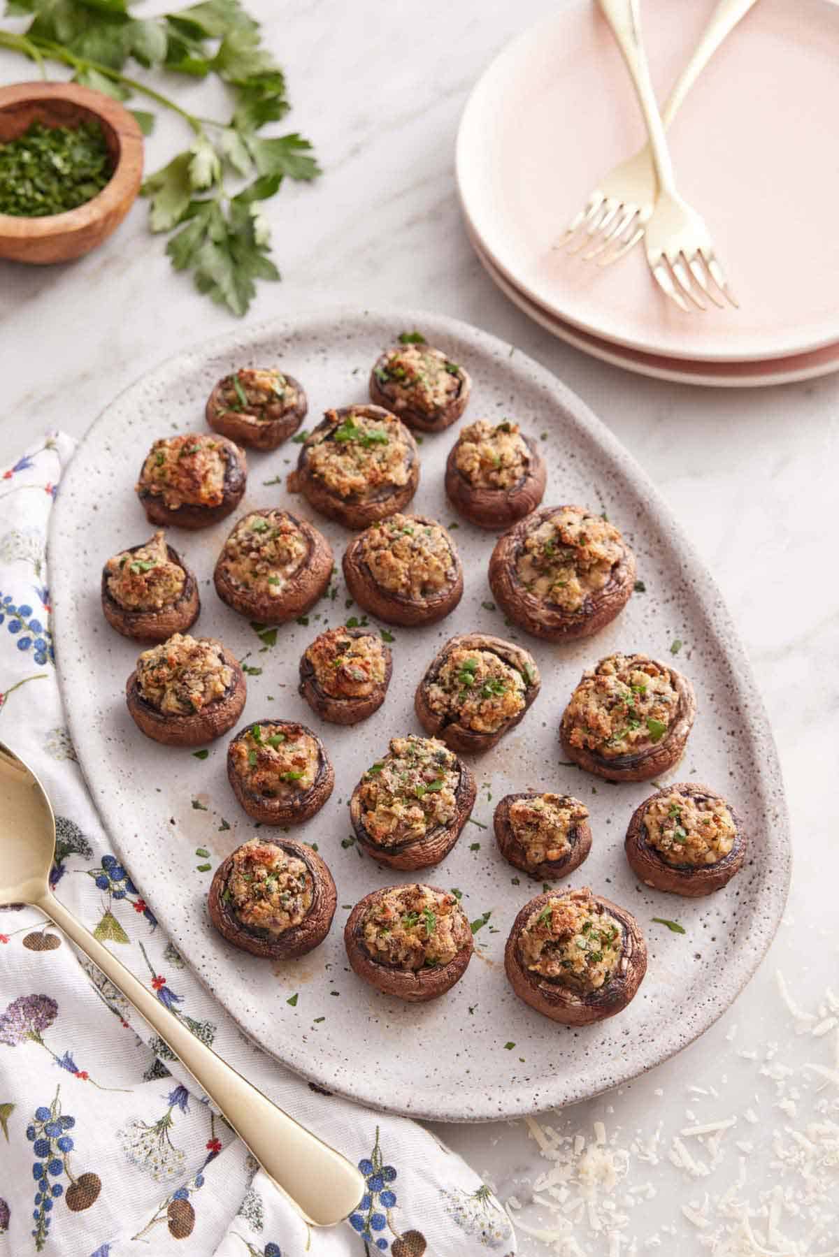 Overhead view of an oval platter of stuffed mushrooms with stacked plates, forks, parsley, and shredded parmesan around the platter.
