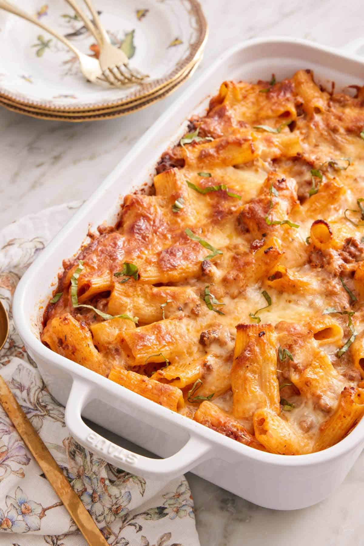 A white baking dish of baked rigatoni with a stack of plates and fork in the back.