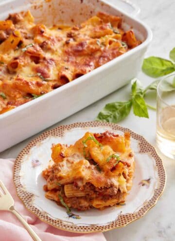 A plate with a serving of baked rigatoni with the baking dish in the back and some basil and wine to the side.