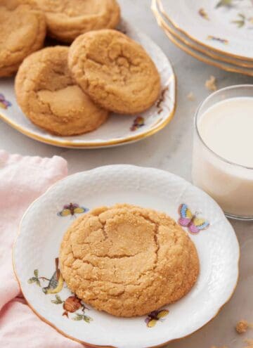 A plate with a brown sugar cookie with a platter with more cookies and a glass of milk behind it.