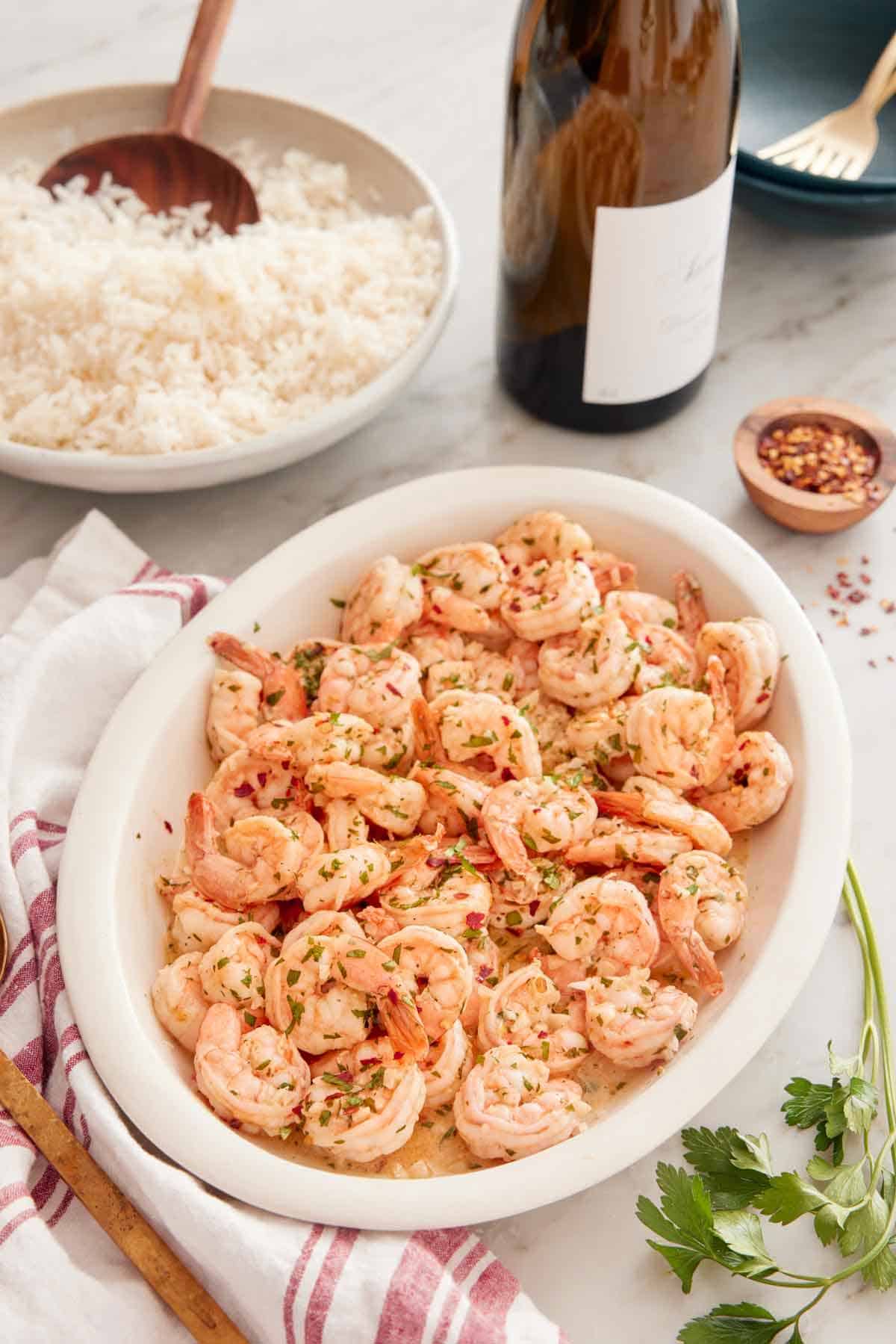 A platter of garlic shrimp with a plate of rice, a bottle of wine, and a pinch bowl of red pepper flakes in the back.