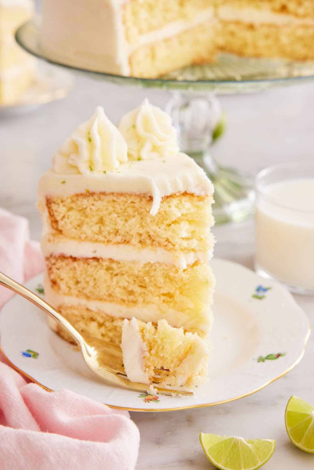 A slice of key lime cake on a plate with a bite on the fork in front. Cut limes in front and glass of milk and cake in the background.