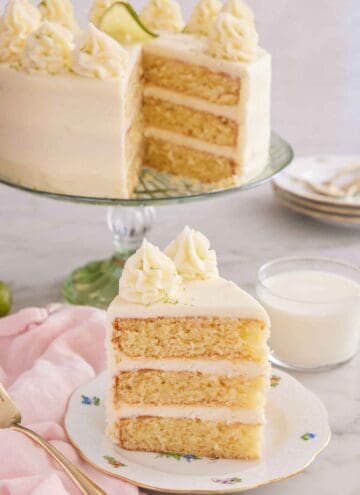 A slice of key lime cake on a plate with a glass of milk and the rest of the cake in the background.