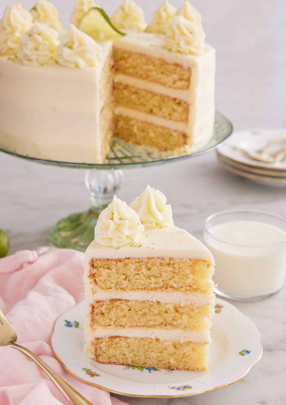 A slice of key lime cake on a plate with a glass of milk and the rest of the cake in the background.
