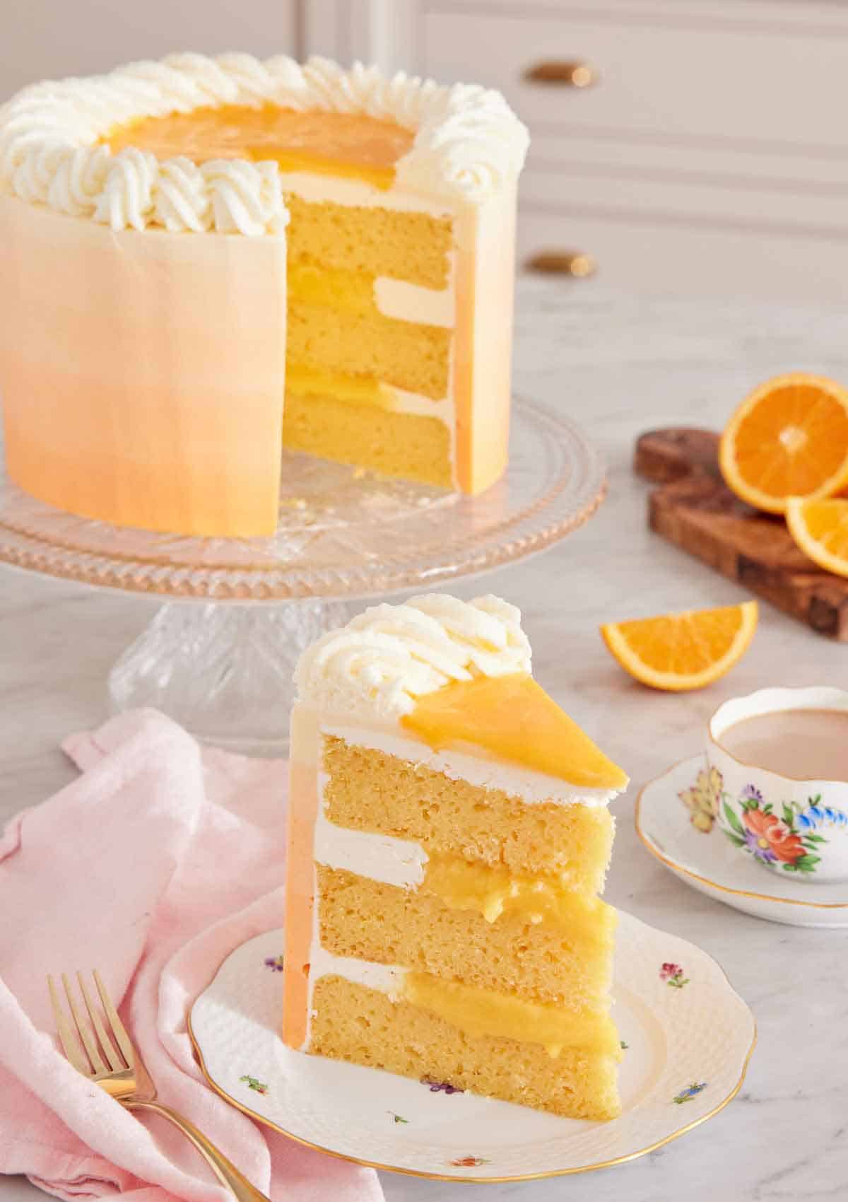 A slice of orange creamsicle cake with the rest of the cake on a cake stand in the background along with some cut oranges and a cup of coffee.