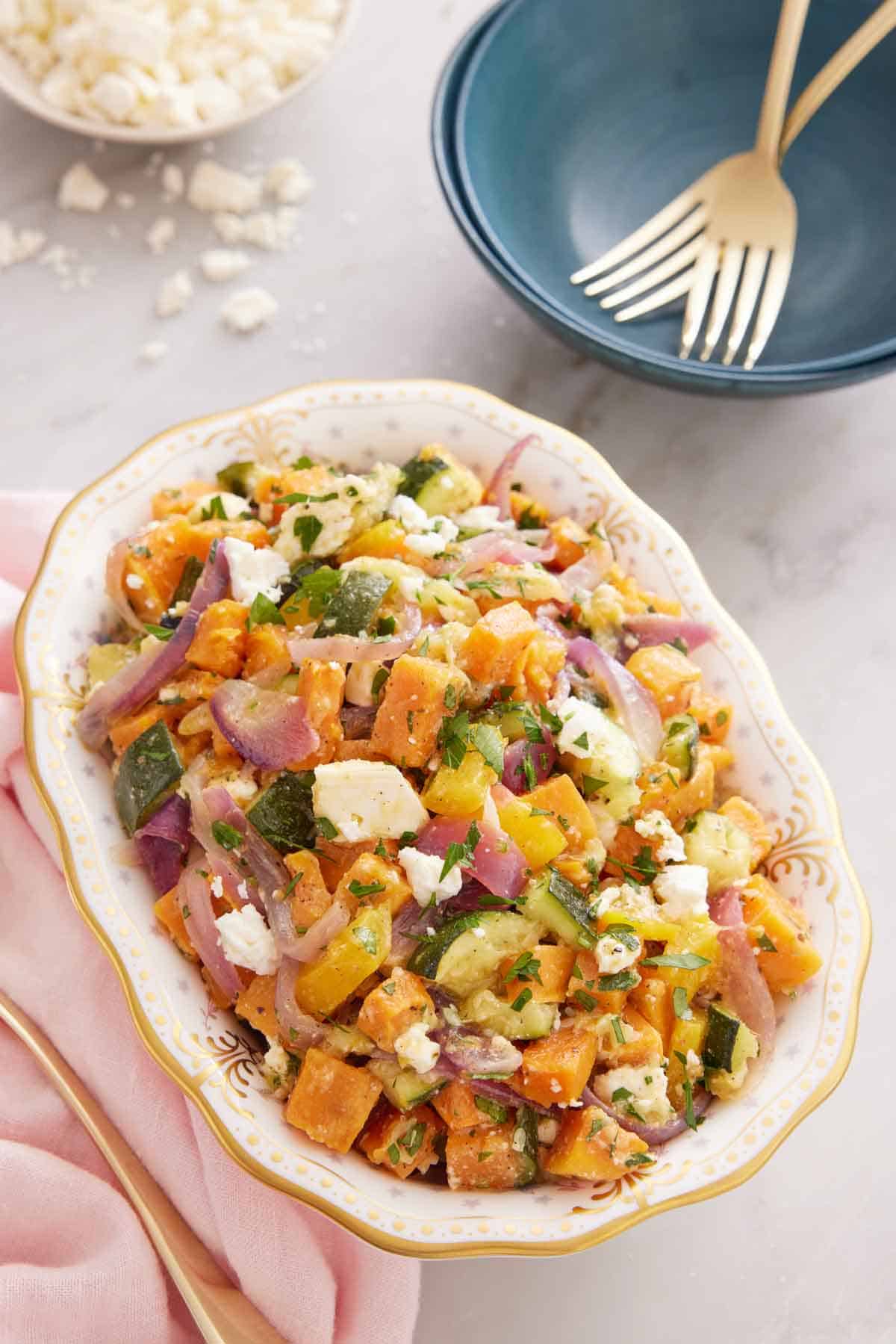 A platter of roasted vegetable salad with a bowl of crumbled feta in the back alongside a stack of bowls and forks.