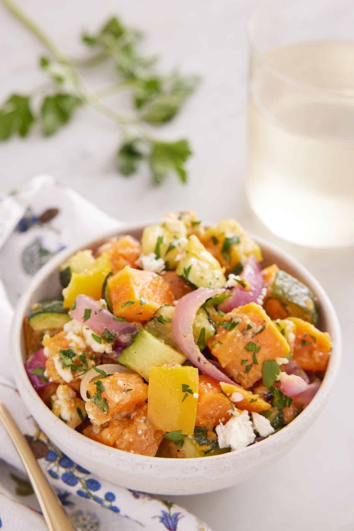 A bowl of roasted vegetable salad with a glass of wine and parsley in the background.