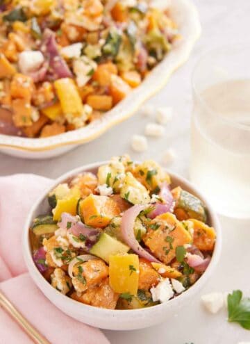 A bowl of roasted vegetable salad with a glass of wine and platter in the back.