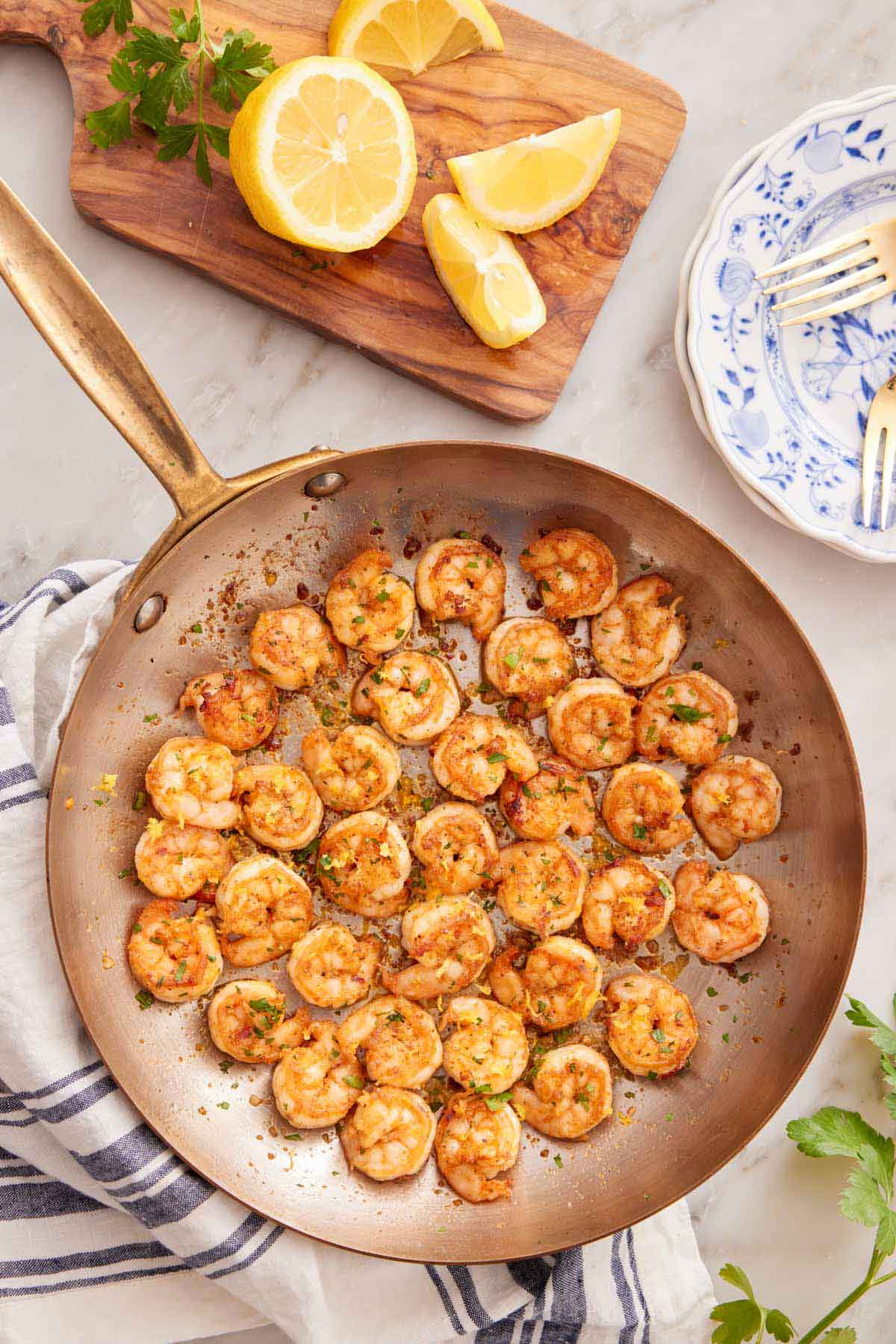 Overhead view of a skillet of sautéed shrimp with a cutting board with lemons and parsley beside it.