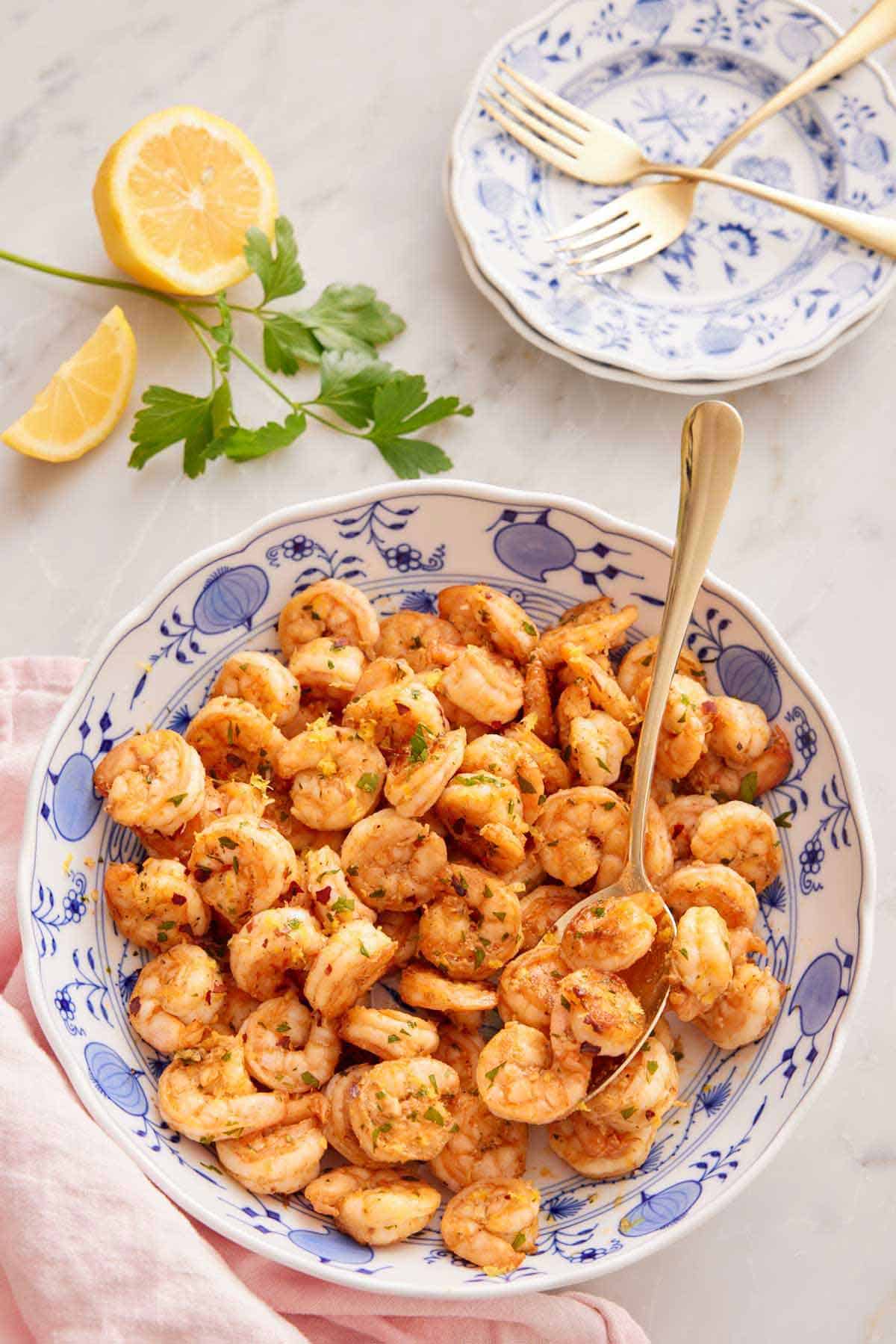 A large serving plate of sautéed shrimp with a spoon with some parsley, lemon, stack of plates, and forks in the back.