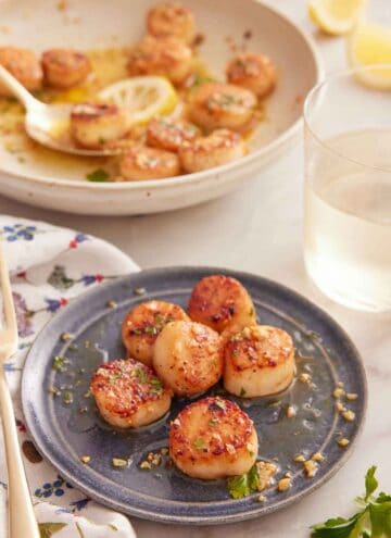 A plate with a serving of seared scallops with a glass of wine and the rest of the scallops in the background.
