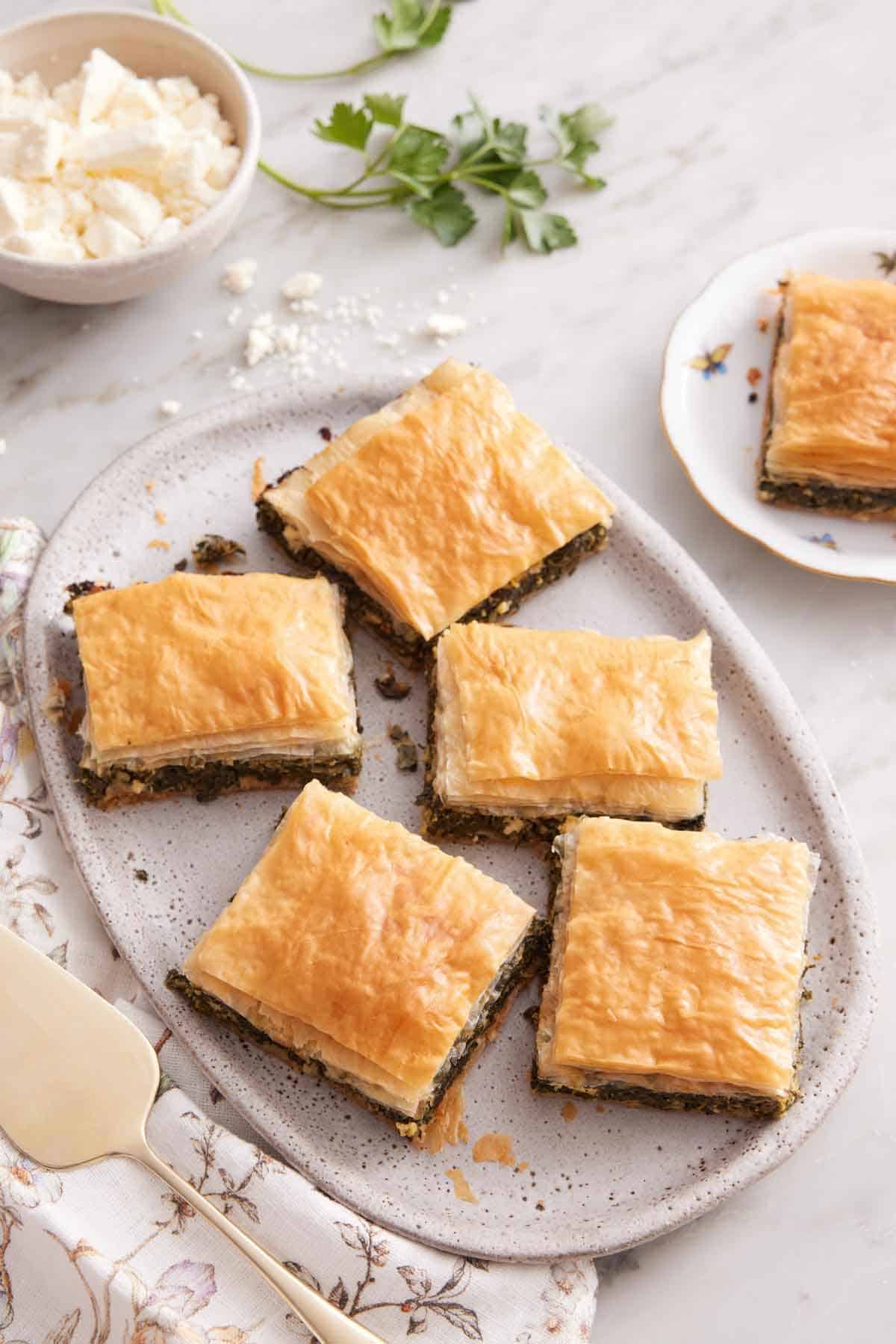 A platter with 5 pieces of cut spanakopita with another plate with a slice on the side.