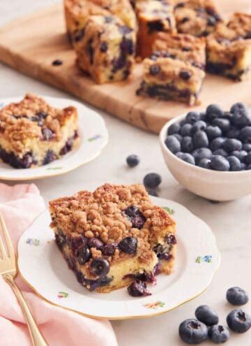 A plate with a square serving of blueberry buckle with a bowl of blueberries and additional cut pieces in the background.