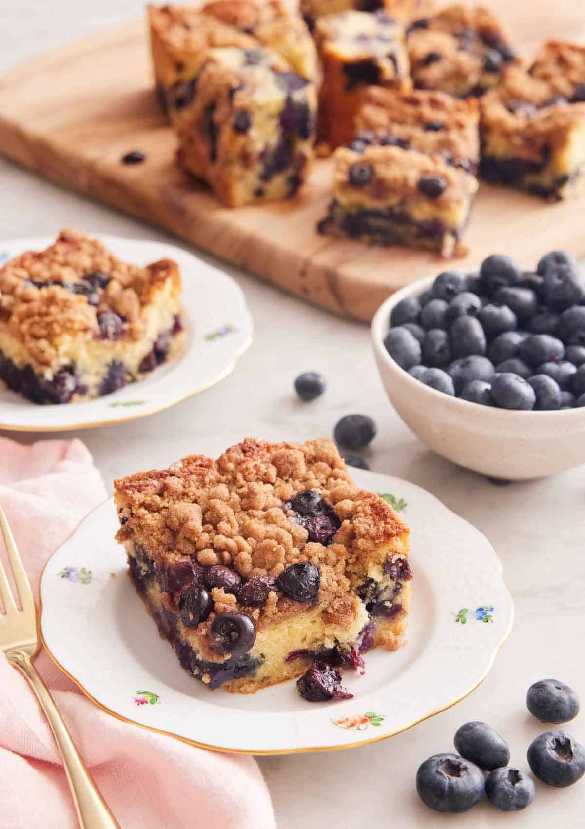 A plate with a square serving of blueberry buckle with a bowl of blueberries and additional cut pieces in the background.