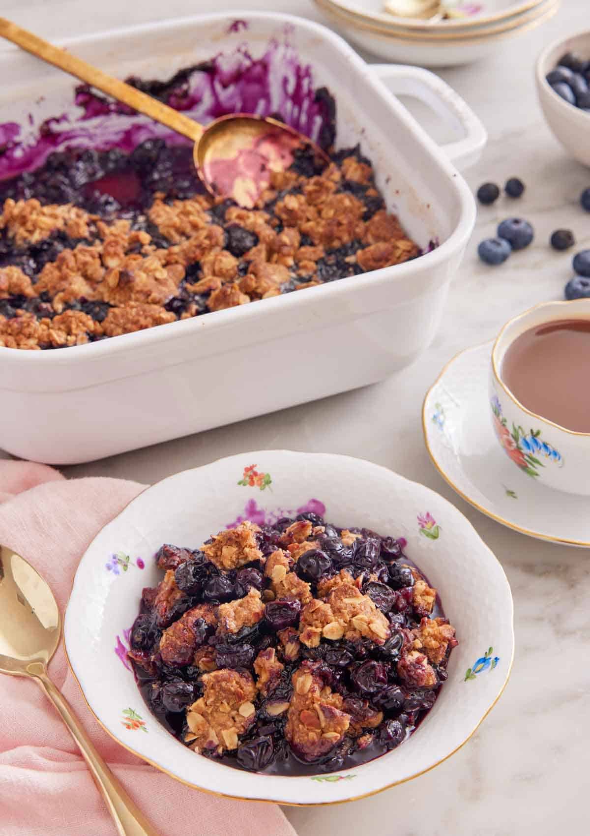 A bowl of blueberry crisp with a casserole dish with the rest of the dessert in the background also with a mug of tea.