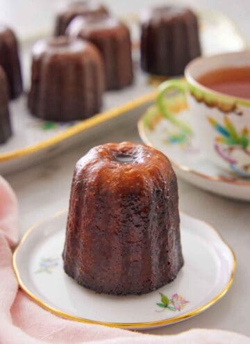 A canelé on a plate with more in a platter in the back along with a cup of tea.