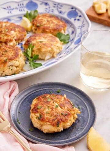 A small plate with a crab cake with a platter with more behind it and glass of wine.