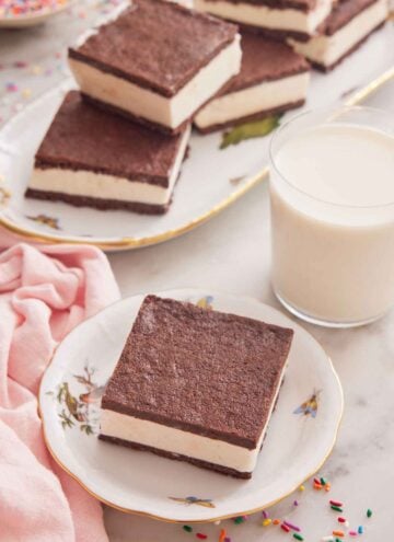 A small plate with a square piece of ice cream sandwich with a glass of milk and additional sandwiches on a platter behind it.