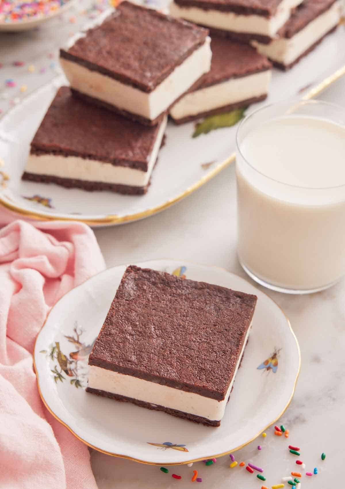 A small plate with a square piece of ice cream sandwich with a glass of milk and additional sandwiches on a platter behind it.